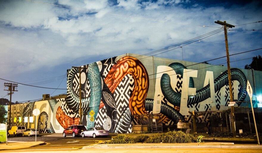 A wide building with a slight industrial appearance, with a mural showing large multicolored snake shapes wrapping around it. Several cars are parked nearby, with a couple of people standing outside the building. There is a dark blue sky above.