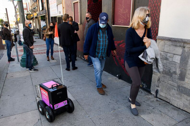A small, pink, four-wheeled delivery robot rolls along a sidewalk next to pedestrians.