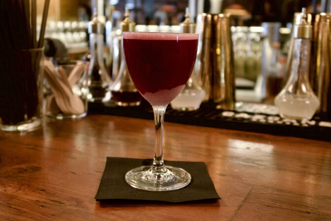 A deep red cocktail in a stemmed glass sits on a bar counter, with bottles and bar tools blurred in the background.