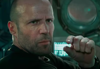 Jason Statham sits in a submarine, the video controls visible behind him. He wears an all back water-suit. He looks off at someone out of frame. His eyes slightly hooded by his eyebrows pressed together, giving a serious look. He holds up one hand his index and thumb in a pinch gesturing to the size of something perhaps. 