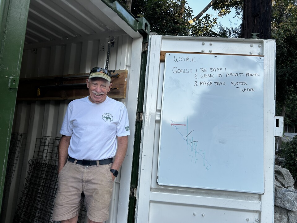An older white man wears a white t-shirt and beige shorts and beige 