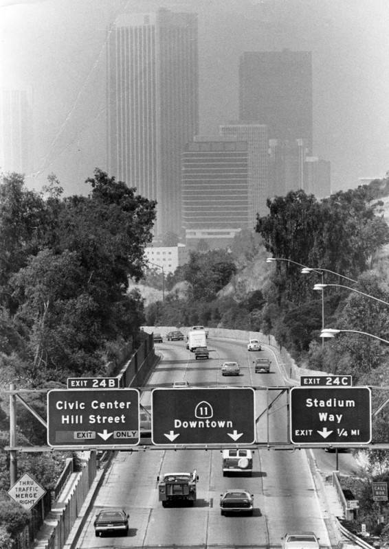 Tall buildings in a black-and-white photograph shrouded in smog with a four-lane freeway and freeway exit signs below.
