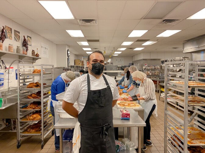 A man wearing a black apron and black face mask stands in the middle of a commercial kitchen. He poses for the camera with his hands behind his back as several people work at a large food-prep table behind him.