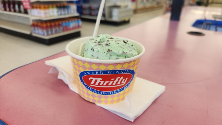 A close up of a cup of ice cream, which says Thrifty in white letters on a red background. Inside there is a green chocolate chip mint icecream, with a plastic spoon. The cup is on a counter in a Rite Aid.