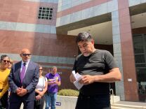 A Latino man in a black T-shirt and pants opens a piece of papaer while other look on outside a building.