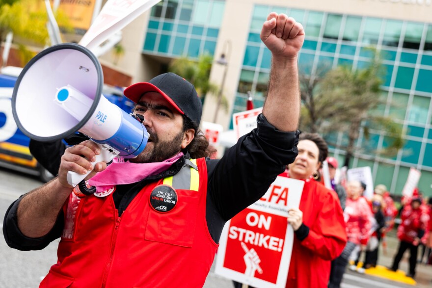 A masculine presenting person with medium-light skin tone wears a red vest over a black shirt and a black and red baseball cap while holding a white and blue megaphone that they hold to their mouth while raising one hand in a fist. Behind the person there's a crowd in red holding signs.
