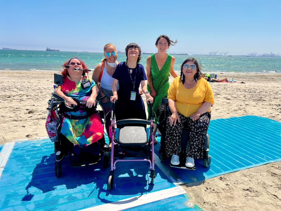 five light skinned people, two in wheelchairs and one with a walker, are standing on a blue plastic beach mat on the sand. Behind them about 30 feet away is the ocean