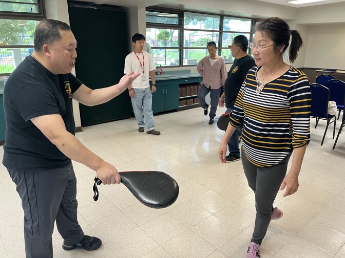 A middle-aged Asian American man holds a paddle up for his student, the middle-aged Asian American woman, to kick.