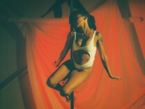 A feminine presenting person with medium-dark skin tone wears black curly hair to the side and a one piece white leotard with the belly cut out while balancing on a pole with their arms to the side. Behind them is a red curtain. 