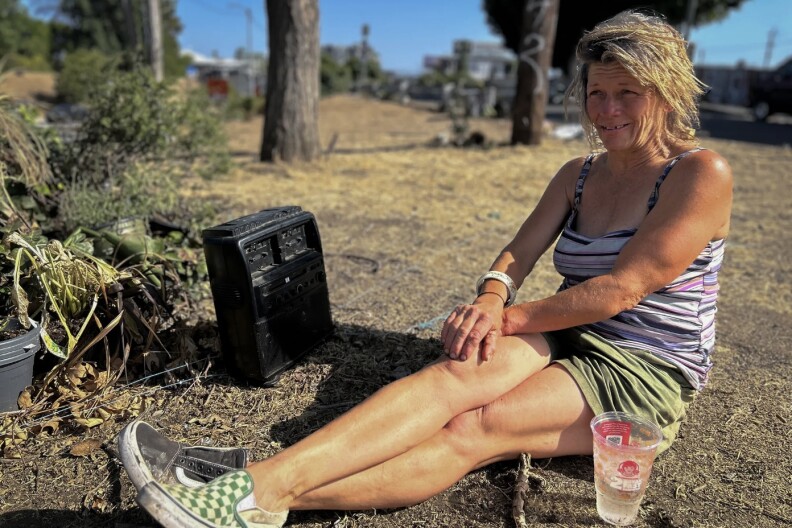A woman with light skin seating on the grass, wearing a purple tank top and green shorts that cross her legs and arms, with an old black radio on her side.