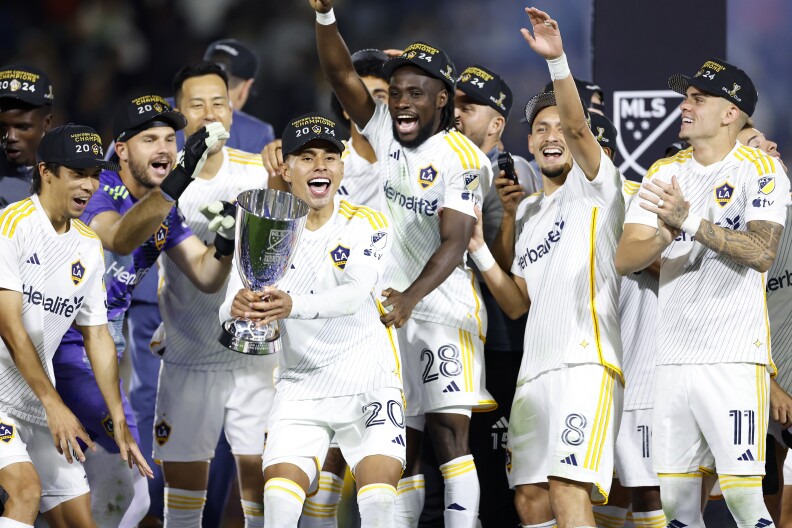 Players on the LA Galaxy soccer team cheer on stage as one player hold the conference trophy in the center of the frame.