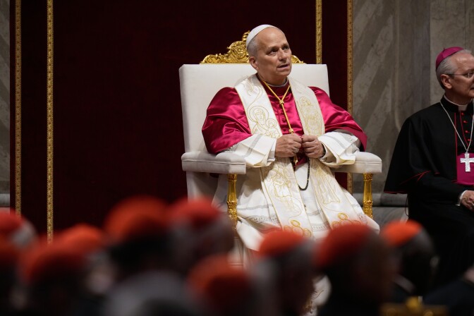 The pope, in a red robe and white stole, sits on a throne-like chair.