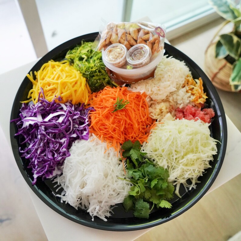 An overhead image of a black bowl that's filled with piles of different types of food, such as white noodles, purple cabbage, green cilantro, light green cabbage and orange carrots.