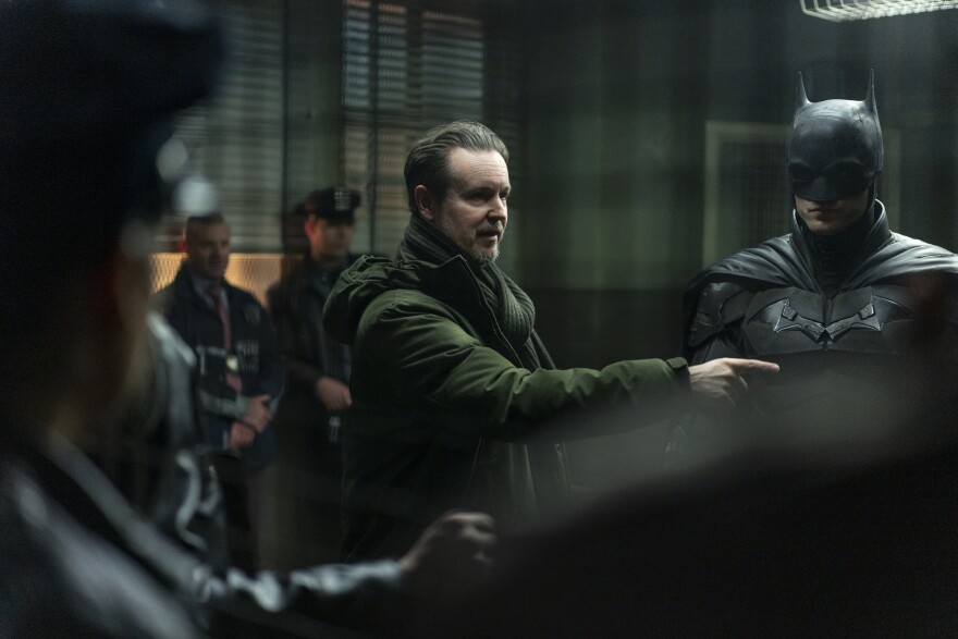 A man in a thick coat with a scarf talks and points, while another man in full Batman costume stands by.