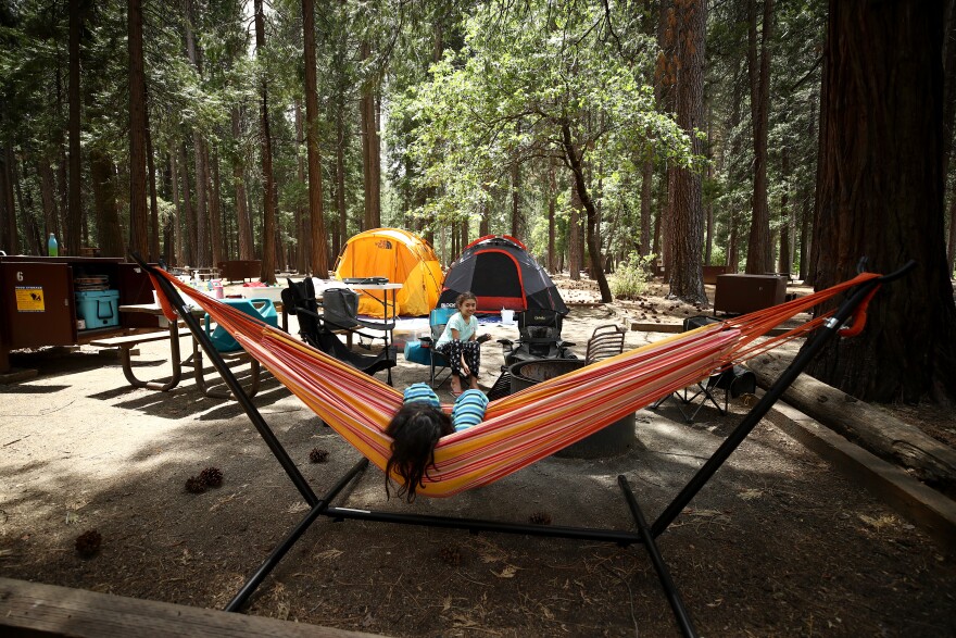 A person relaxes in a colorful hammock in a forest while a child sits next to a firepit. A couple of tents are behind them.