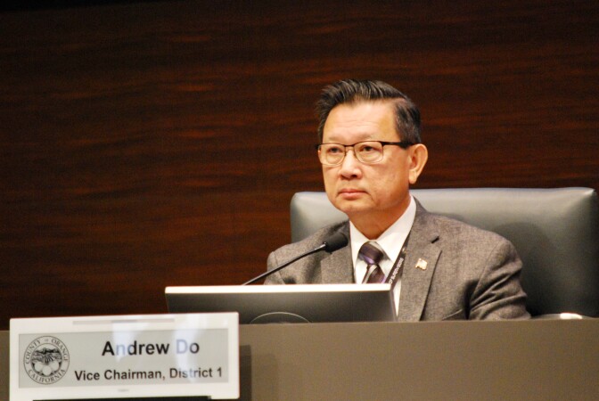 A man in a chair wearing a suit jacket, tie and glasses looks forward with a microphone in front of him. A sign in front has the official seal of the County of Orange and states "Andrew Do, Vice Chairman, District 1."
