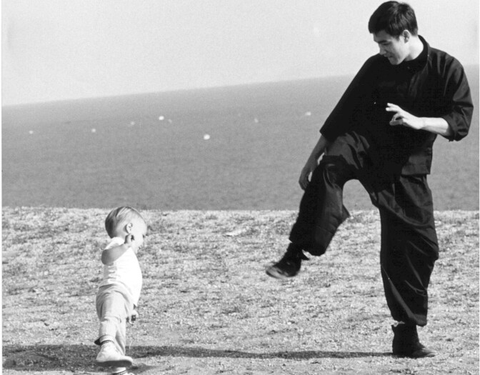 A smiling Asian American man in a black gi and his son play on a grassy bluff by the ocean.   