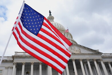 An American flag waving in the foreground, with the U.S. Capitol dome visible in the background under a partly cloudy sky.