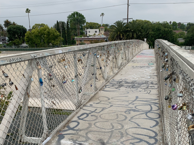 A narrow pedestrian bridge that is marked up with graffiti is lined by white metal railings and fencings on both sides. People have taken to attacking padlocks, of all different sizes and colors, onto the fencing as a sign of their devotion to someone else.