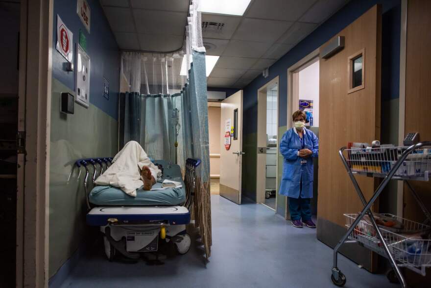 A patient on a gurney in a hospital corridor waits for a room. A curtain provides some privacy. In the background, a nurse wearing protective gear stands in a doorway.