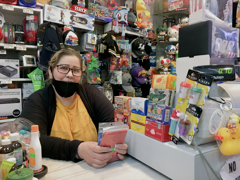 A woman in a black mask and yellow shirt at the register of a small store, surrounded by various products.