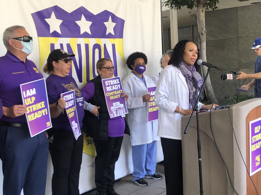 A group of peope stand behind the podium. One person is at the mic speaking. The others are behind her holding signs with statements such as "I am ULP strike ready."