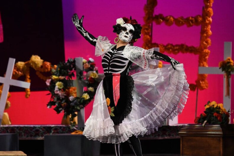Image is a woman on stage performing while wearing a calacas mask and a skeleton halloween type of costume. Behind her is staging for the show.