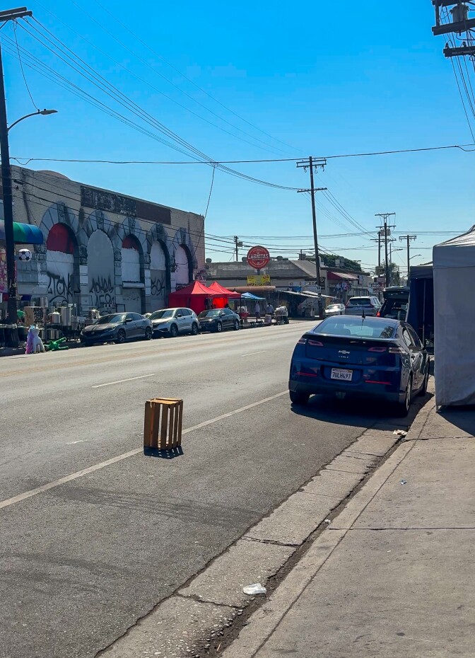 Cars are parked on both sides of a street, a wooden crate behind one of them. Power lines and store signs are visible in the background.