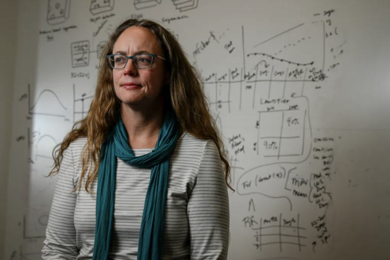 A woman with darkish-blonde hair, light skin and wearing glasses looks off to the left. Behind her is a whiteboard full of scribbles notes and graphs.