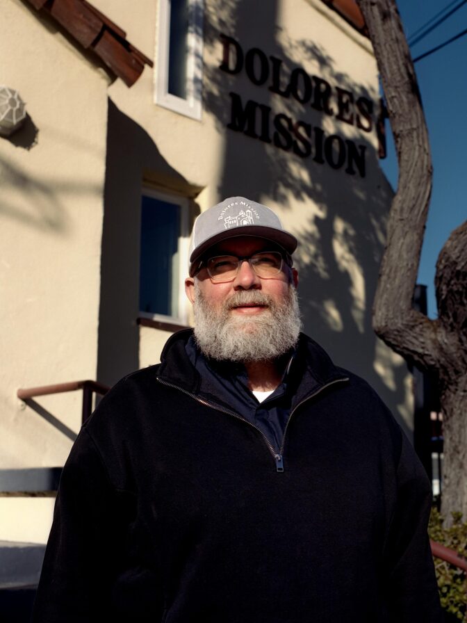 A man with a grey beard, wearing a grey cap, eyeglasses and a black sweater with a zipper.