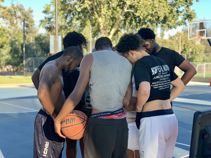 A group of men stand in a huddle on a basketball court, heads down. One man, back to the camera, has a basketball in his left hand.