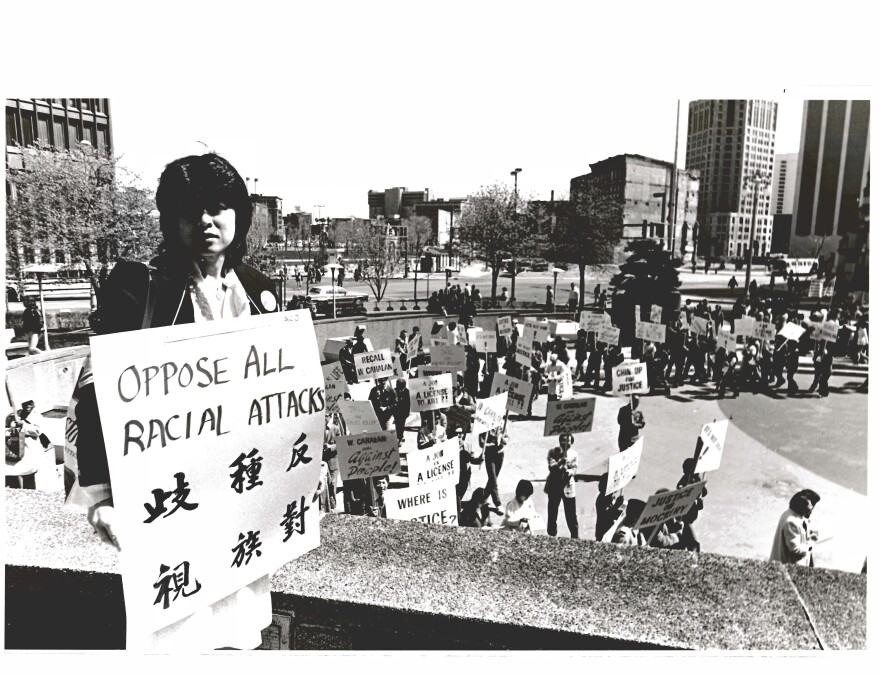 In a black-and-white photo, a person holds a sign reading "Oppose all racial attacks." In the background other protesters are gathered.