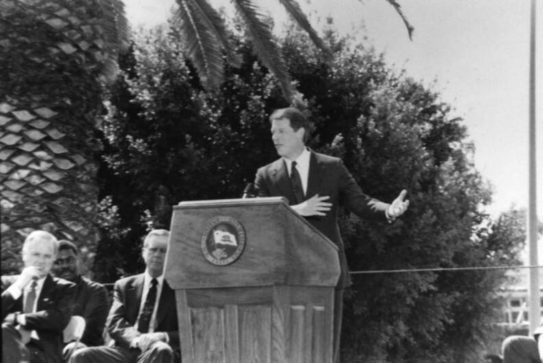 Vice president Al Gore stands at a podium outside with trees behind him. two men are sitting to his right while the vice president is talking.