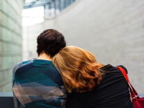 A man and woman are shown from behind. The woman leans her head on the man's shoulder. The woman has shoulder-length red hair and the man has short brown hair. 