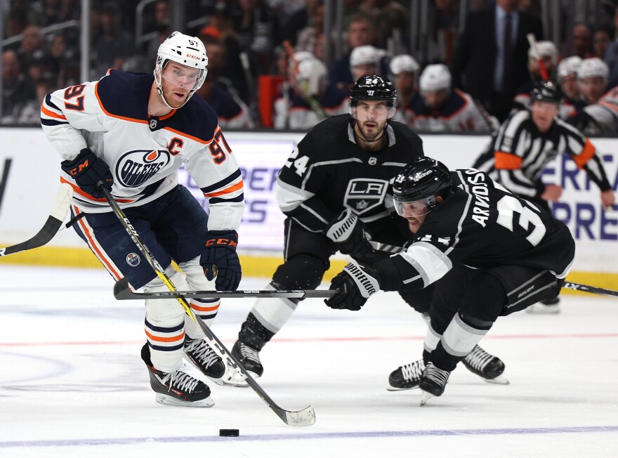 Three hockey players on ice skates, two wairing black jerseys with Los Angeles Kings shield logos with white armbands and white socks and black helmets, move toward a player wearing a white, orange, and blue jersey with and a white helmet in a hockey rink. The player in white, Connor McDavid, pulls ahead of the nearest Kings player, Viktor Arvidsson.