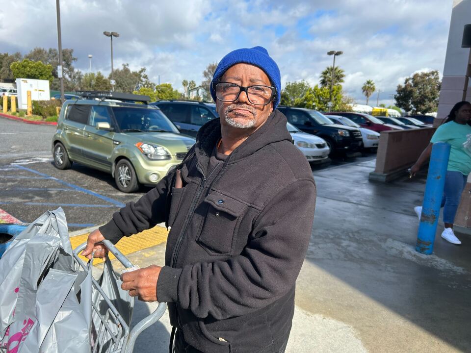 A man wearing glasses and a blue beanie pushing a shopping cart stocked with gray bags of goods in a store parking lot.