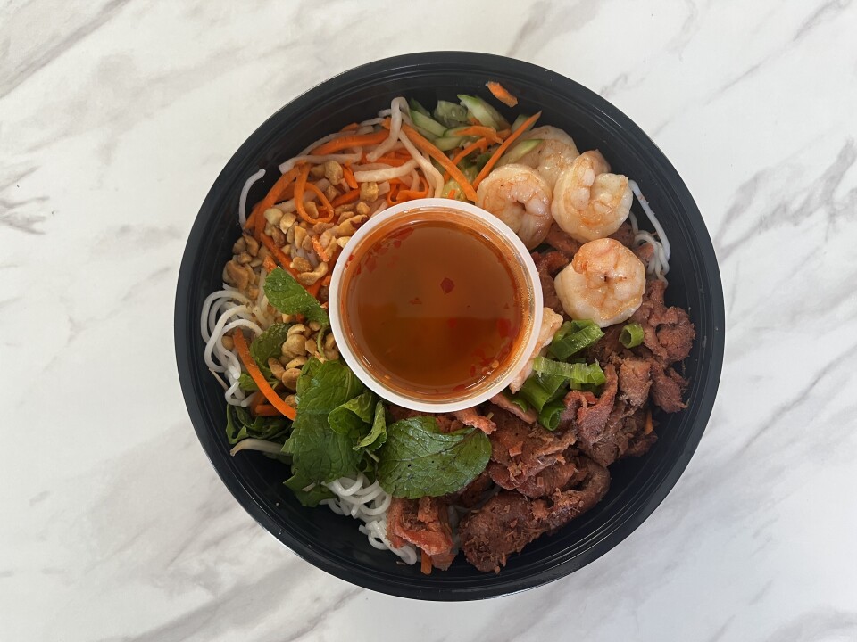 A bowl of thin white noodles that contained grilled meat, small shrimp, and assorted vegetables covered in nuts and mint. In the center is a plastic container containing an orange dressing.  
