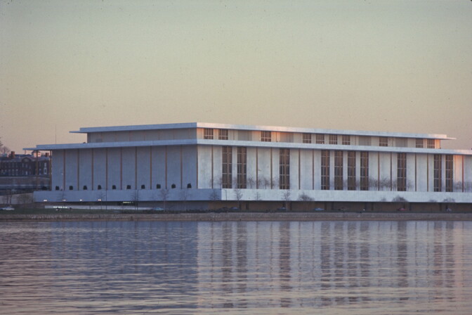A large, low white building sits on the bank of a river. It is pictured from across the river.