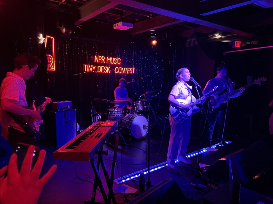 A woman playing guitar and standing in spotlight before a microphone leads a band that includes two other guitarists and a drummer. They are on a darkened stage with an illuminated sign behind them that reads NPR TINY DESK CONCERT.
