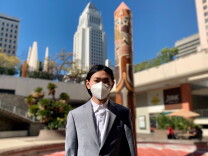 A young Chinese man poses in a gray suit with Los Angeles City Hall in the background. 