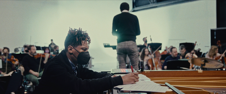 A man in a black coat wearing a medical mask composes music atop a piano. A conductor and studio musicians holding instruments can be seen in the background.