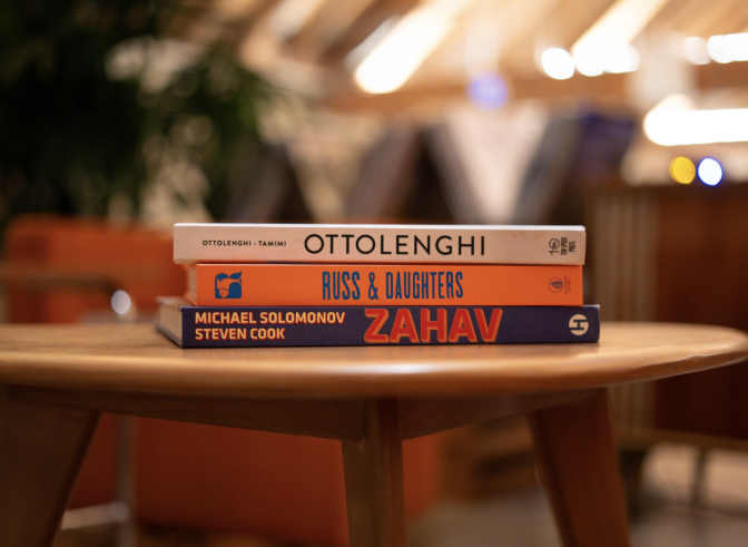 Three books are piled on top of each other on a wooden table; the book titles are shown on the spines: Ottolenghi, Russ and daughters and Zahav