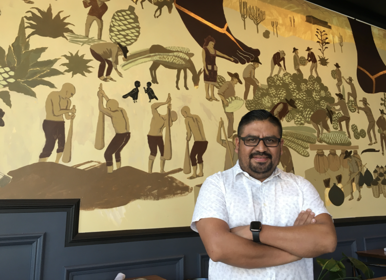 A brown-skinned man in a white shirt stands in front of a mural in tones of yellow and brown.