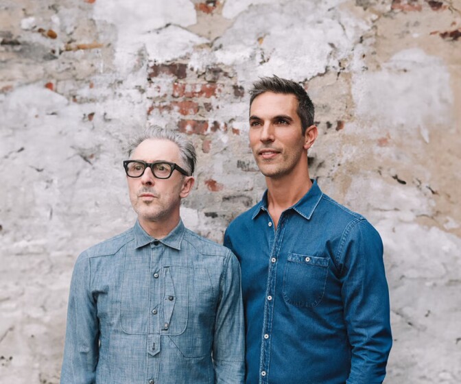 Alan Cumming (left) and Ari Shapiro stand together in front of a brick and plaster wall, both wearing denim and looking off into the distance. 