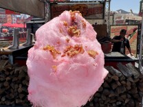 A hand is holding up a stick of pink cotton candy that is sprinkled with bacon bits. 