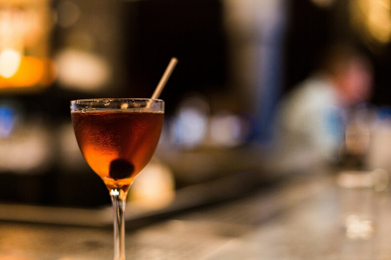A small cocktail glass filled with an orange-brown liquid and a dark cherry at the bottom, with a toothpick sticking out over the edge.