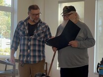 Two men stand and look at a binder containing a work of William Shakespeare. There is a window behind them and some wooden chairs. 
