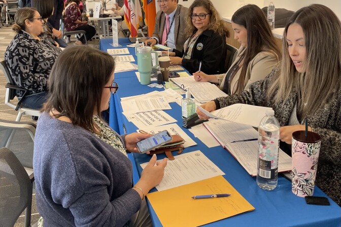 Seven people sit on both sides of a table draped by a blue tablecloth and covered with paperwork.