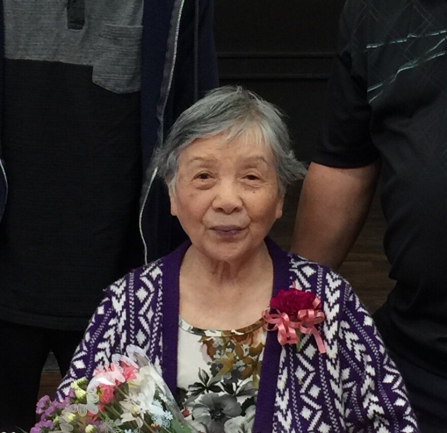 an image of 93-year-old Tomiko Nakayama, 93, who had hoped to stay at her senior home in Boyle Heights for the rest of her life. She has short black-and-gray hair and she wears a sweater with a corsage as she also holds a bunch of flowers.