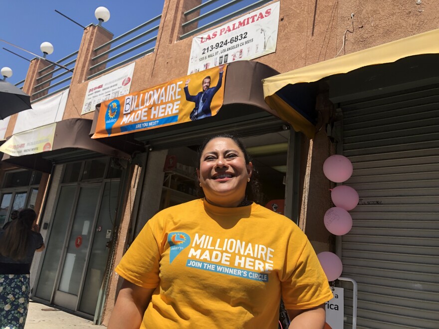 A woman with light brown skin stands in front of a store. She wears a bright yellow t-shirt that says "millionaires made here."